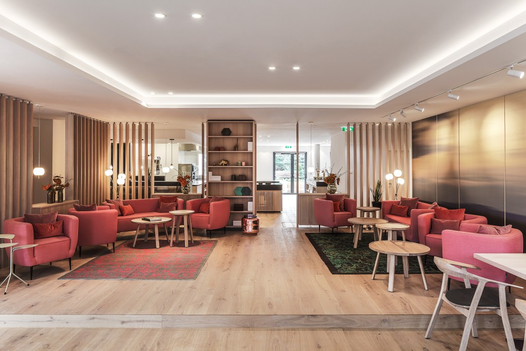 Moderne Innenansicht eines stilvollen Raumes mit rosa Sofas, kleinen Tischen und Holzdetails, gut beleuchtet mit natürlichem Licht.